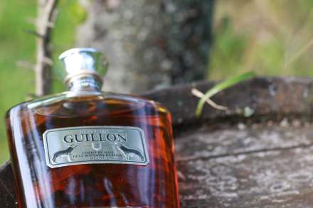 Bouteille de whisky Guillon posée sur une surface en bois avec étiquette argentée, en extérieur près d’un arbre – Fayet Hospitality
