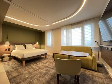Chambre moderne avec lit double, canapé vert et bureau, éclairage design et grandes fenêtres, Fayet Hospitality.