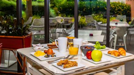 Table de petit-déjeuner bien garnie avec œuf à la coque, brouillade, viennoiseries, fruits frais, céréales, jus d’orange et café, installée devant une baie vitrée donnant sur une terrasse verdoyante.