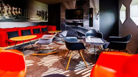 Espace lounge de l’Hôtel Ibis Reims Tinqueux, avec grand canapé rouge, fauteuils design, tables basses et décoration moderne autour d’un écran de télévision.