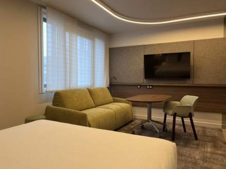 Chambre élégante avec canapé vert, table ronde, fauteuil et télévision murale, éclairage moderne, Fayet Hospitality.