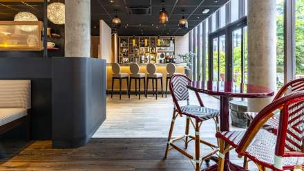 Espace bar élégant avec comptoir lumineux, tabourets design et table bistro rouge près des baies vitrées, Fayet Hospitality.