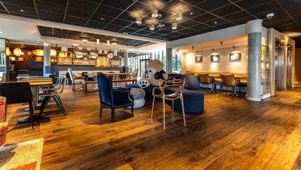Lobby spacieux et chaleureux avec parquet en bois, fauteuils design, tables basses, coin bar éclairé et un grand ours en peluche posé sur un canapé au centre de la pièce.