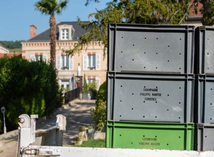 Pile de caisses grises et vertes marquées Champagne Philippe Martin Cumières sur un véhicule, avec maison bourgeoise et jardin en arrière-plan, Fayet Hospitality.