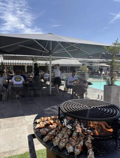 Barbecue avec brochettes grillées au premier plan, terrasse ombragée avec tables près d’une piscine, Fayet Hospitality.