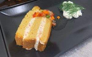 Tranche de cake salé garnie de crème et petits légumes colorés, présentée sur une assiette noire avec herbes fraîches en décoration – Fayet Hospitality