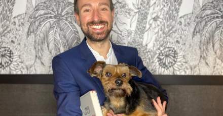 Petit chien à poil court et brun posé sur les genoux d’une personne vêtue d’une veste bleue, fond avec motif floral en noir et blanc – Fayet Hospitality