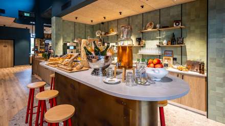 Espace petit-déjeuner du greet hôtel Reims Tinqueux, avec comptoir central présentant viennoiseries, baguettes, boissons, fruits frais et décoration chaleureuse aux tons bois et verts.