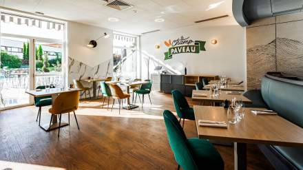 Salle lumineuse du restaurant Le Champ Paveau au Novotel Reims Tinqueux, avec tables en bois dressées, chaises vertes et jaunes, grandes fenêtres donnant sur la terrasse et la piscine.
