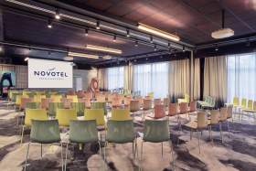 Salle de conférence du Novotel aménagée en théâtre, avec rangées de chaises colorées face à un écran de présentation.